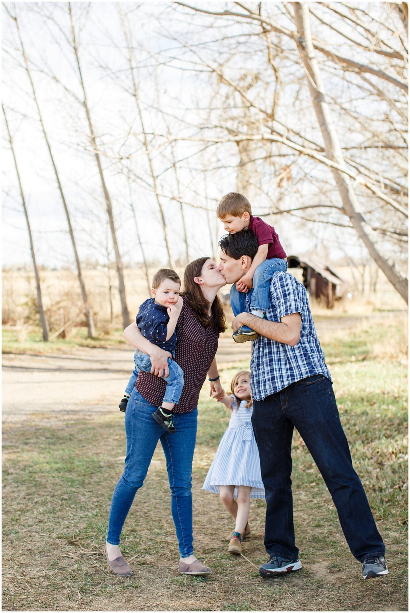 A mother and father kiss as they each hold onto a son and their daughter during an outdoor family photo session