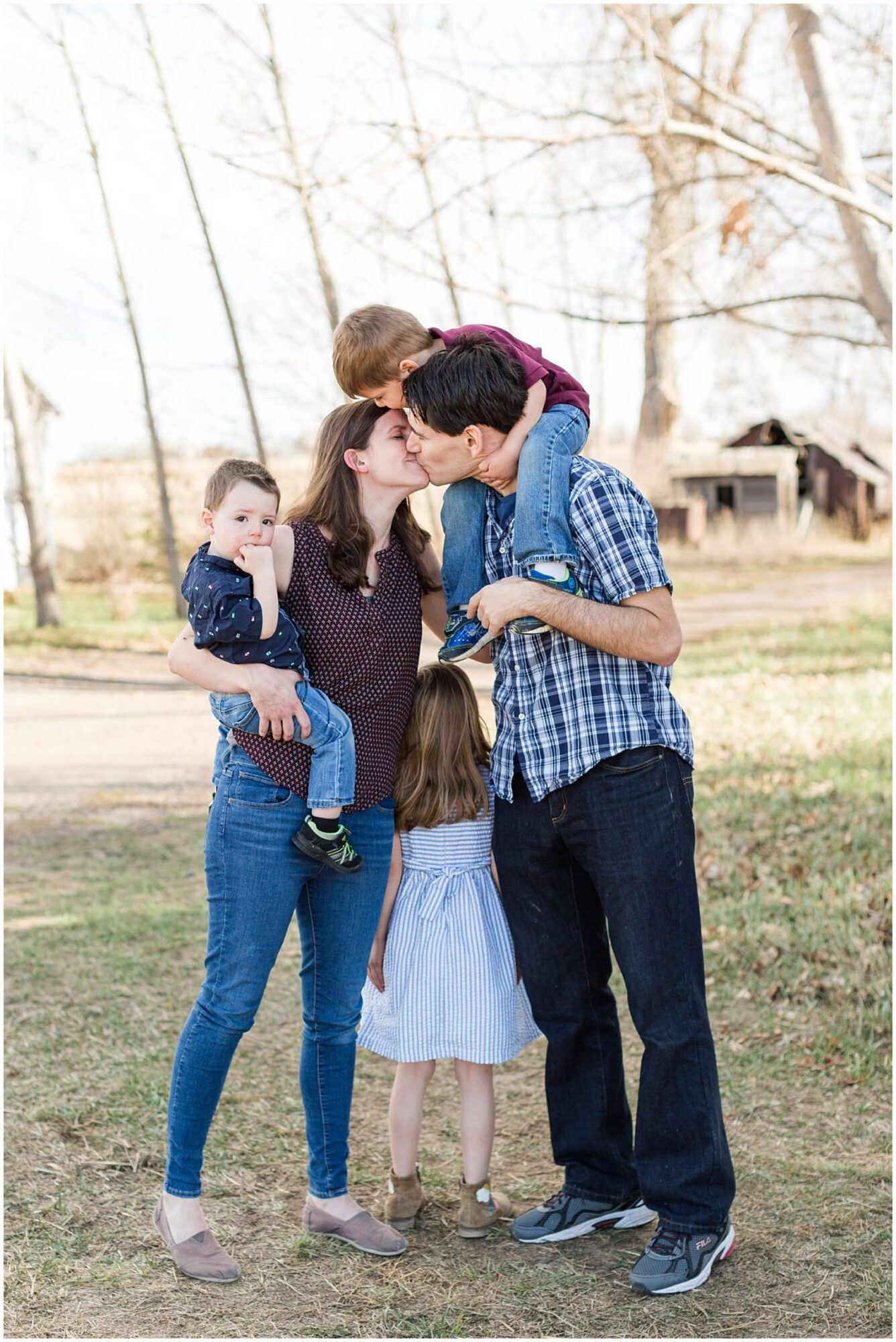 A young husband and wife share a kiss as they stand with their daughter between them and hold a son during an outdoor family portrait session in Erie Colorado