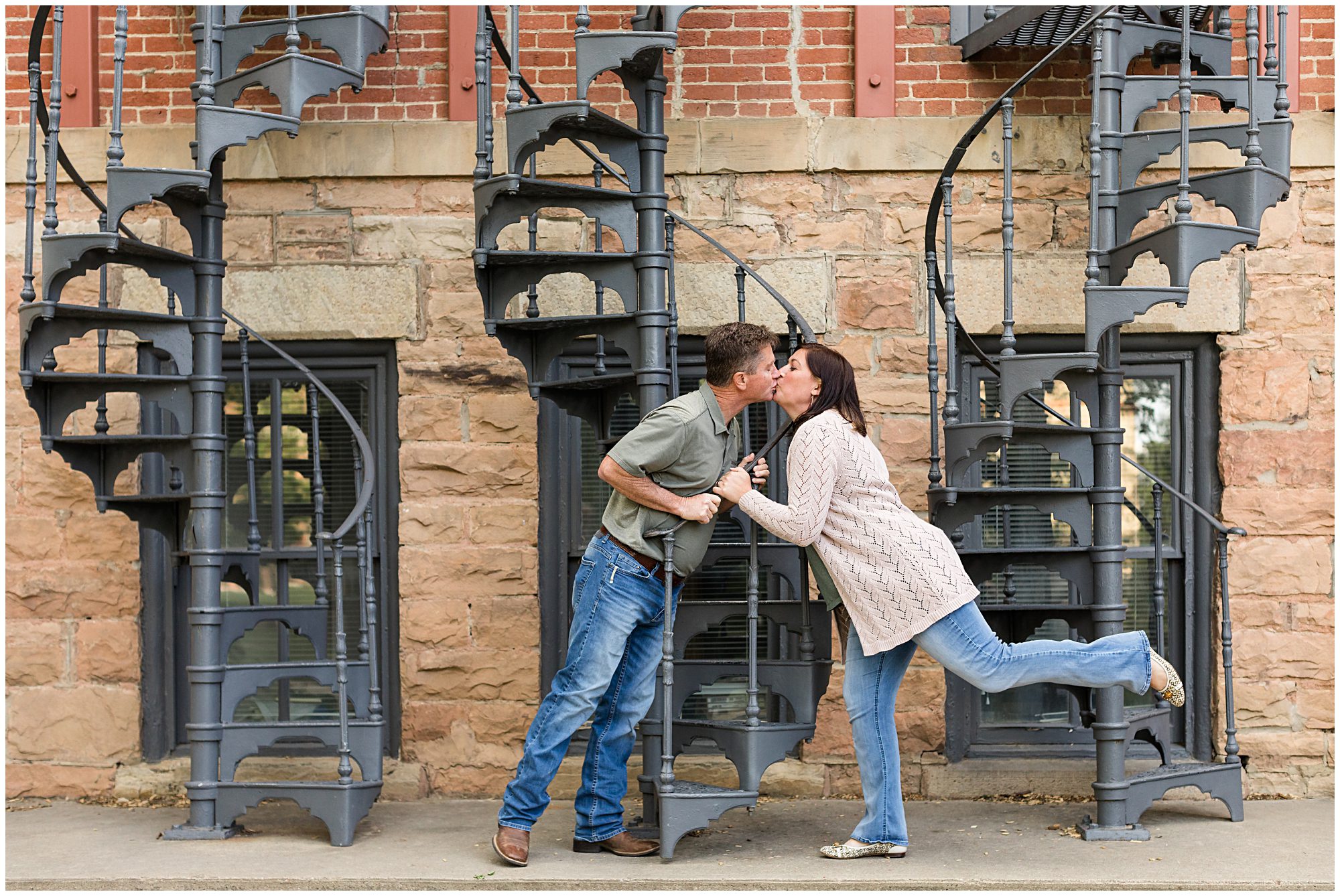 A couple kiss as they lean over the railing of a curved iron staircase during a family photo session in Boulder