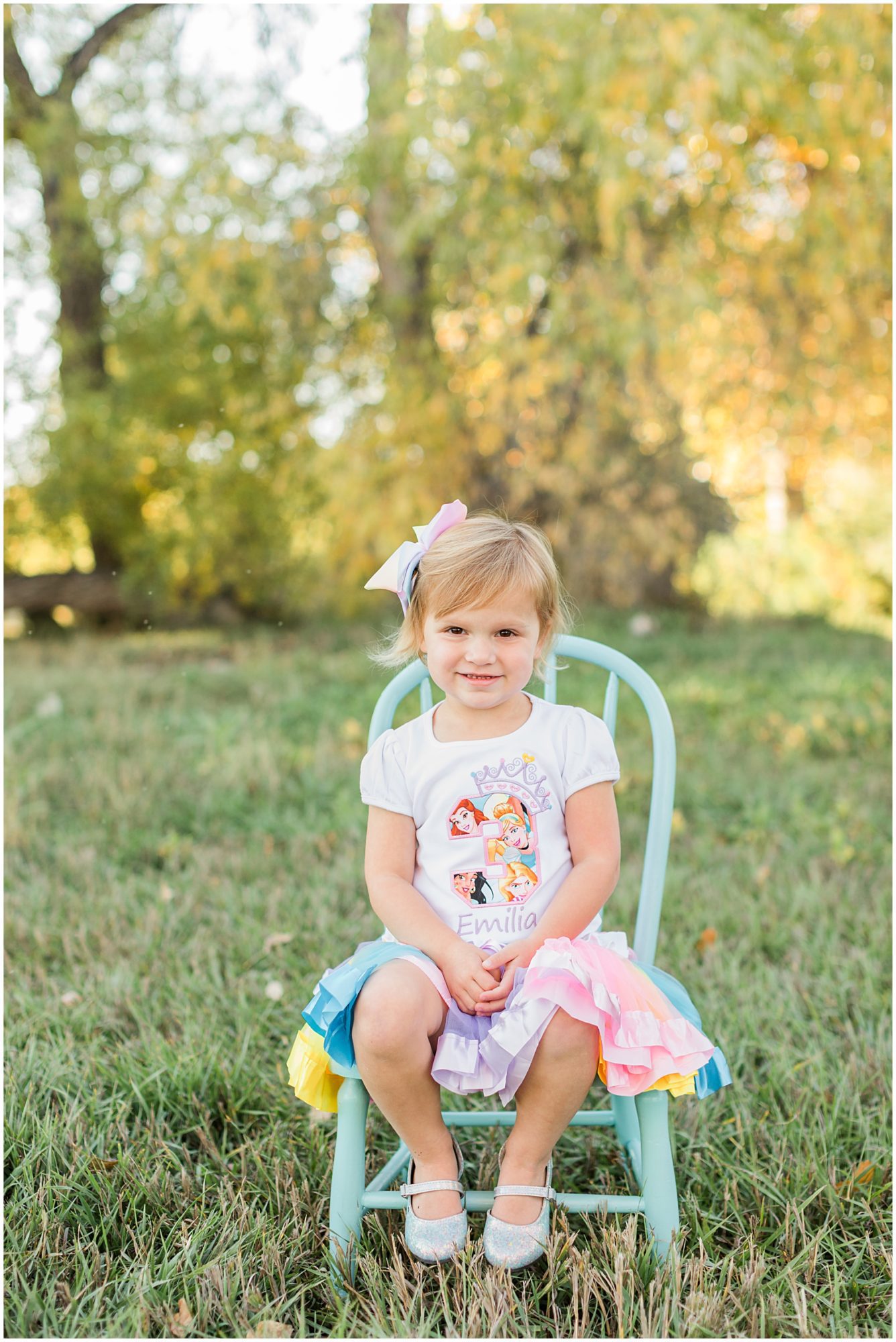 A little girl dressed in a 3rd birthday shirt poses on a blue chair in front of fall colored trees during a mini session at Thomas Reservoir