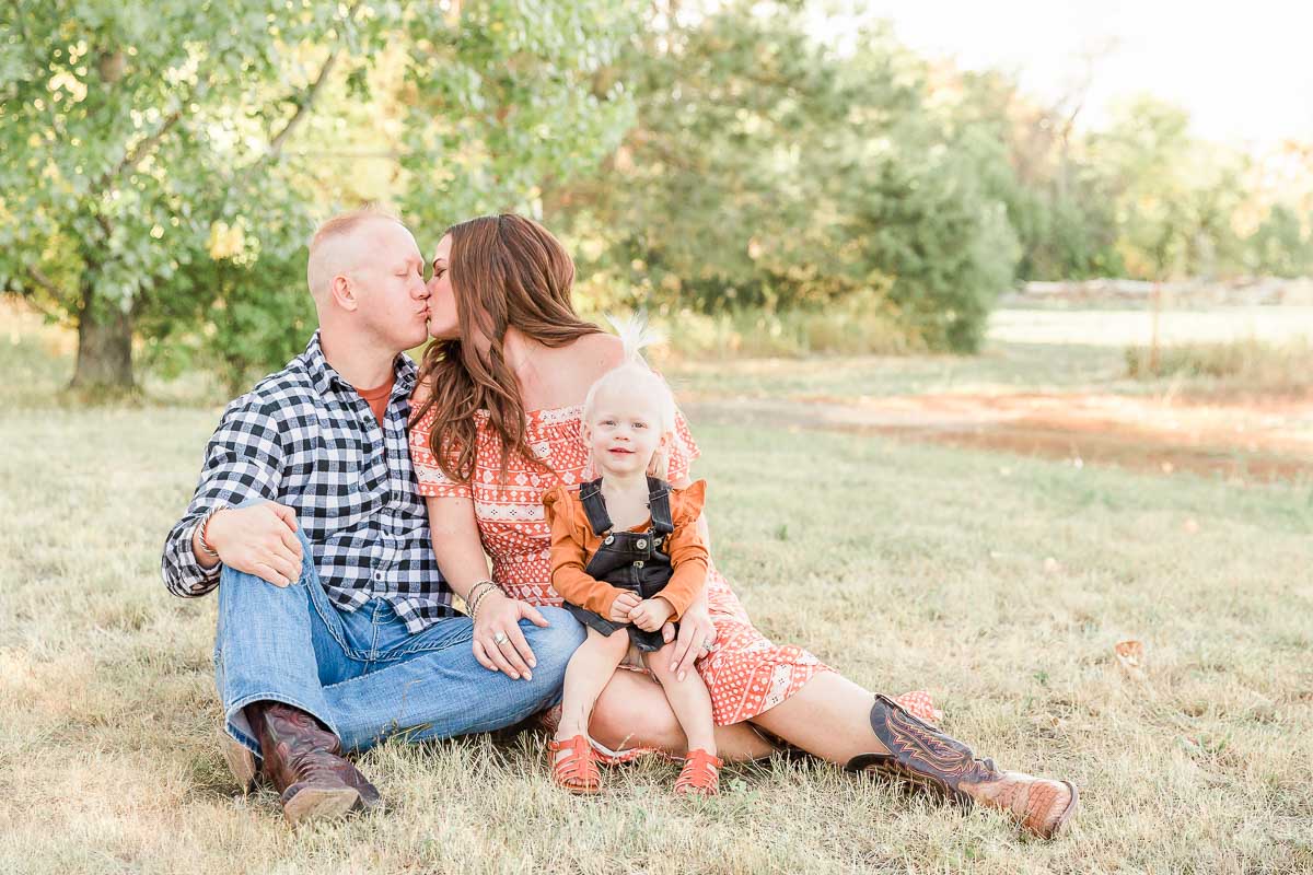 Couple sitting on the grass kissing while their toddler smiles, during a family photo session in Erie, Colorado.