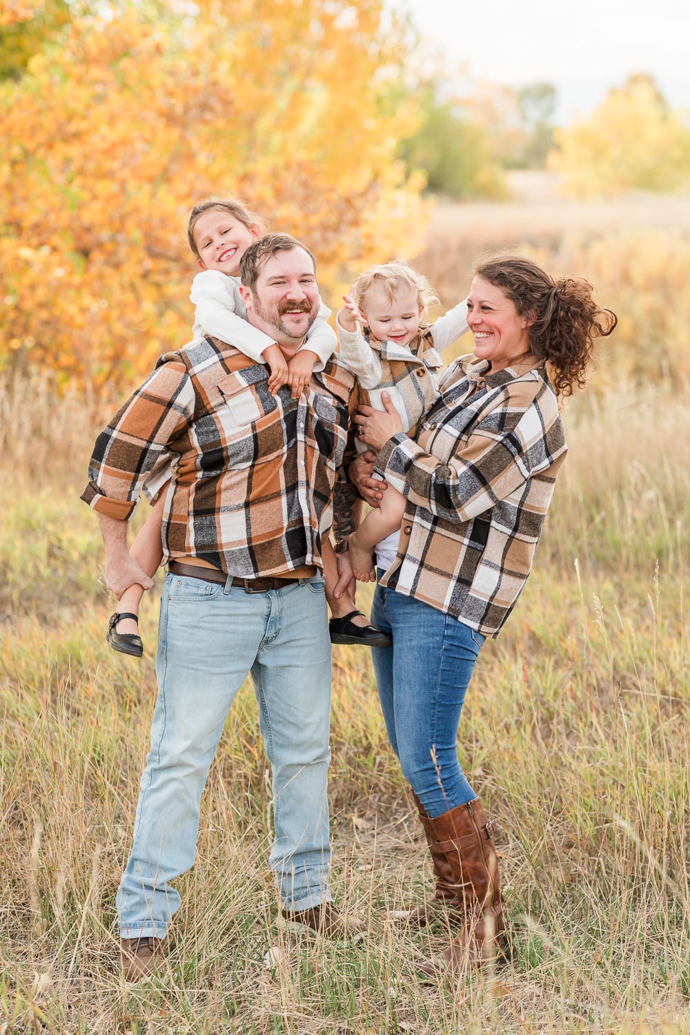 Fall mini session at Coal Creek in Erie, Colorado—young family laughing together in a golden field, wearing matching plaid shirts with autumn trees in the background. Captured by Theresa Pelser, Erie family photographer.