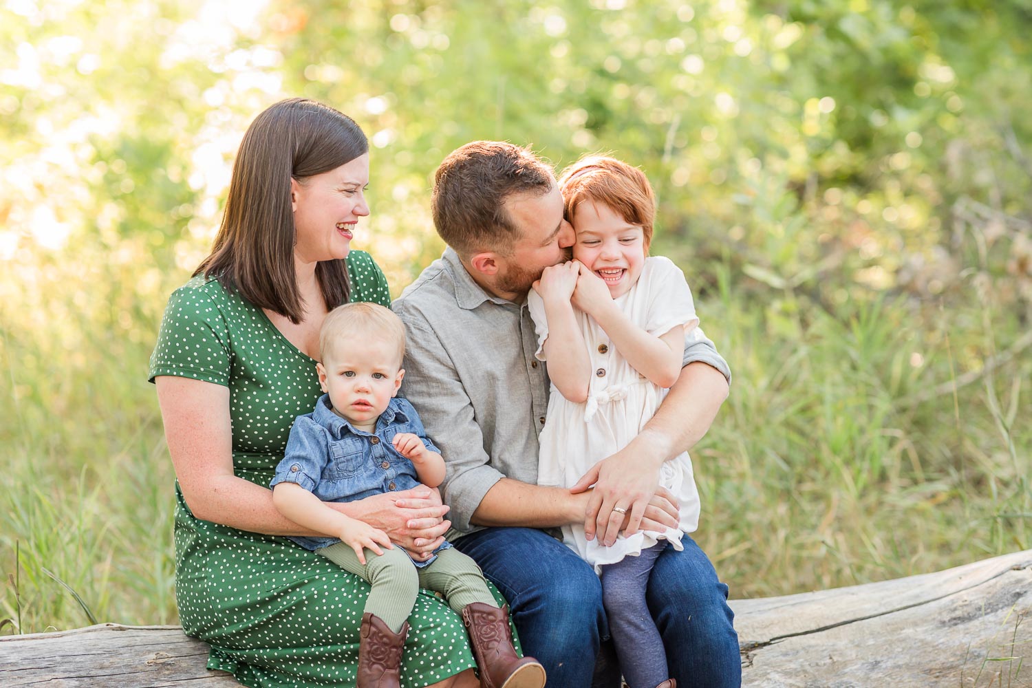 Fall mini session at Coal Creek in Erie, Colorado—parents sitting on a log with their two young children, sharing laughter and snuggles in a sunlit field. Captured by Theresa Pelser, Erie family photographer.