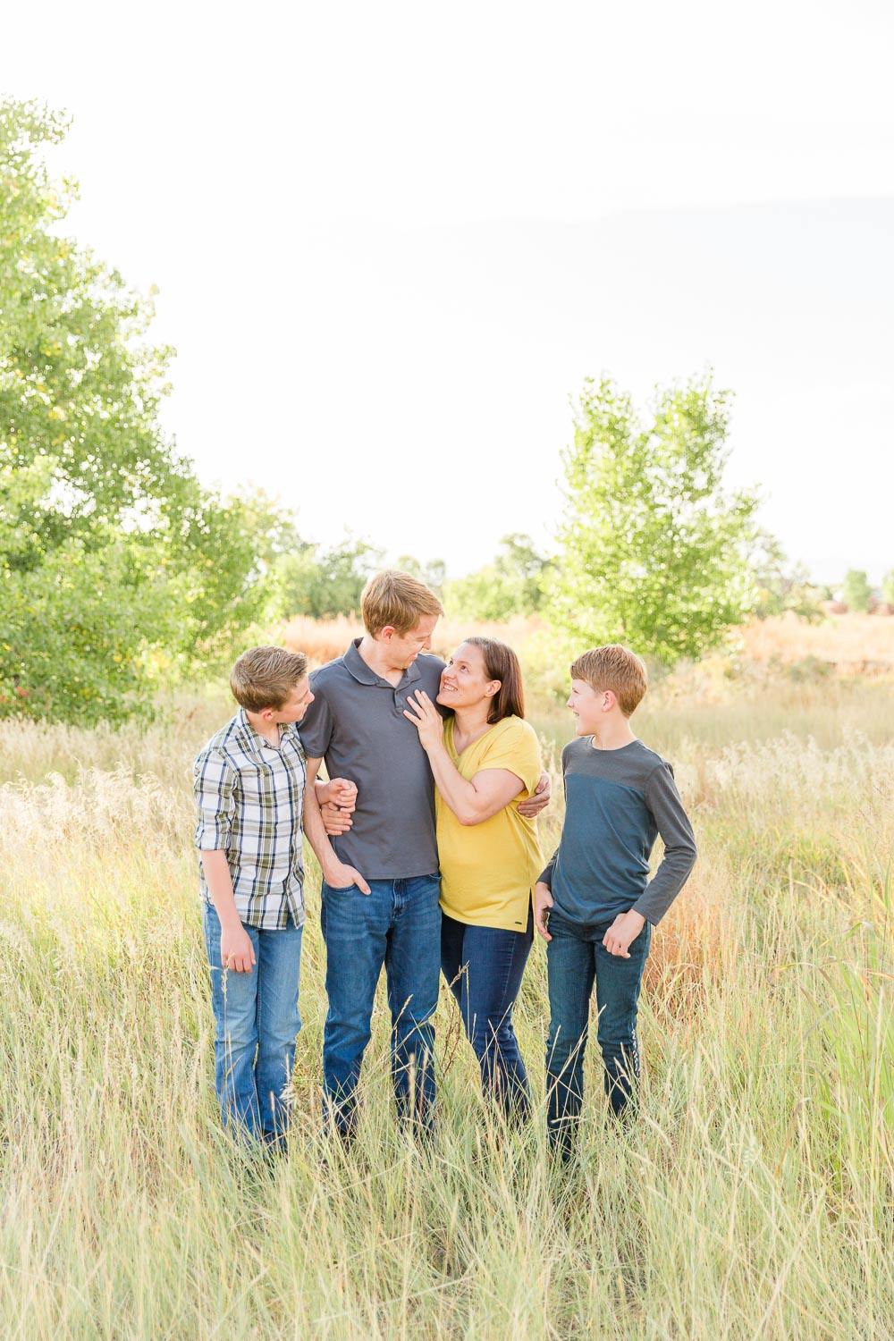 Family mini session at Coal Creek in Erie, Colorado—parents with their two preteen sons standing together in a sunlit field, sharing a warm, candid moment surrounded by tall grass and trees. Captured by Theresa Pelser, Erie family photographer.