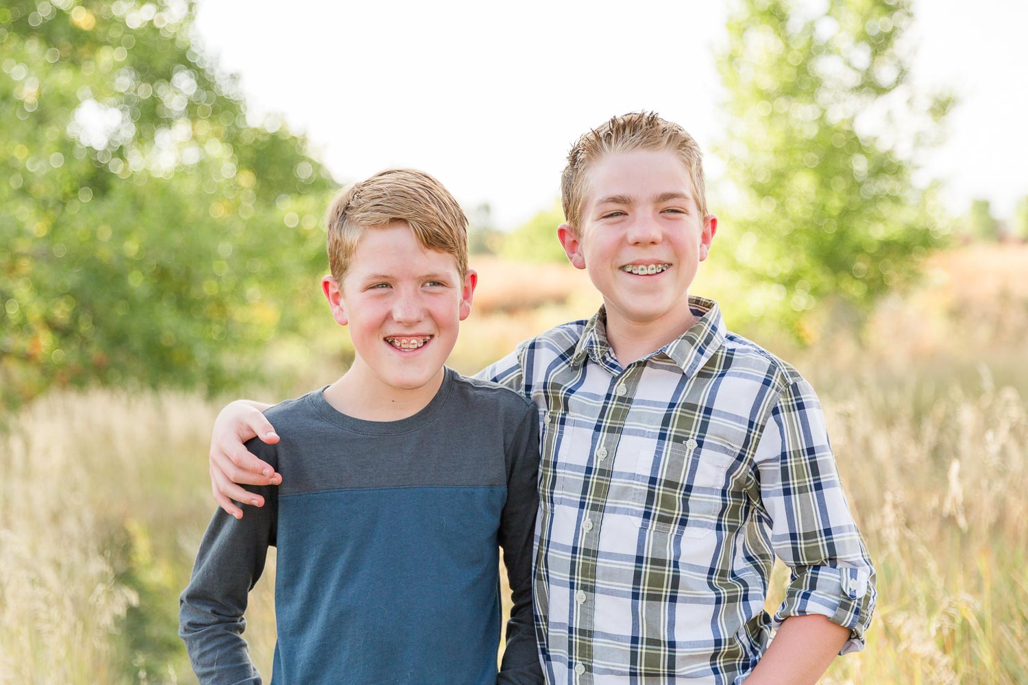 Fall mini session at Coal Creek in Erie, Colorado—two brothers standing close together and smiling in a sunlit field with trees and tall grass in the background. Captured by Theresa Pelser, Erie family photographer.