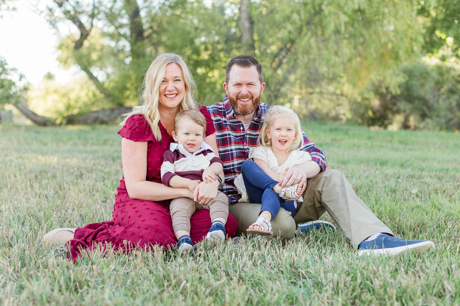 Family of four sitting on the grass during a fall mini session at Thomas Reservoir in Erie, Colorado—mom in a red dress, dad in plaid, with two young kids smiling in a sunlit field. Captured by Theresa Pelser, Erie family photographer.