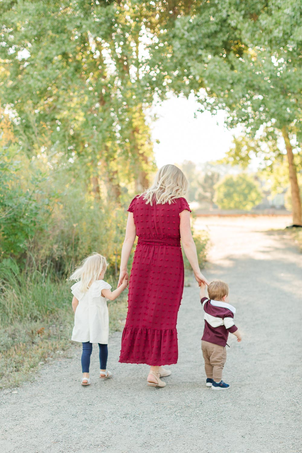 Fall mini session at Thomas Reservoir in Erie, Colorado—mother walking hand-in-hand with her two young children down a sunlit path lined with trees. Captured by Theresa Pelser, Erie family photographer.