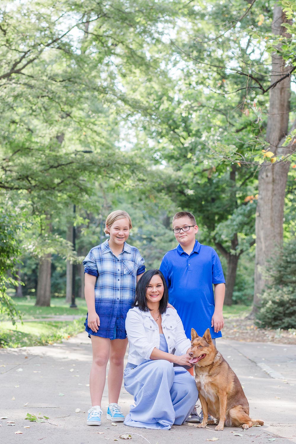 Family session in Boulder, Colorado—mom with her twin son and daughter posing with their dog in a peaceful park setting with tall trees and dappled sunlight. Captured by Theresa Pelser, Boulder family photographer.