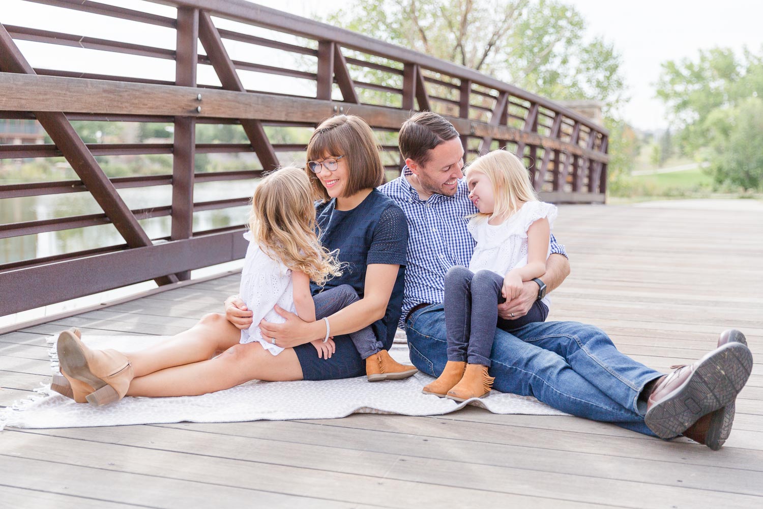 Family session at Anthem in Broomfield, Colorado—parents sitting on a bridge with their two young daughters, sharing a playful and cozy moment during an outdoor photo session. Captured by Theresa Pelser, Broomfield family photographer.