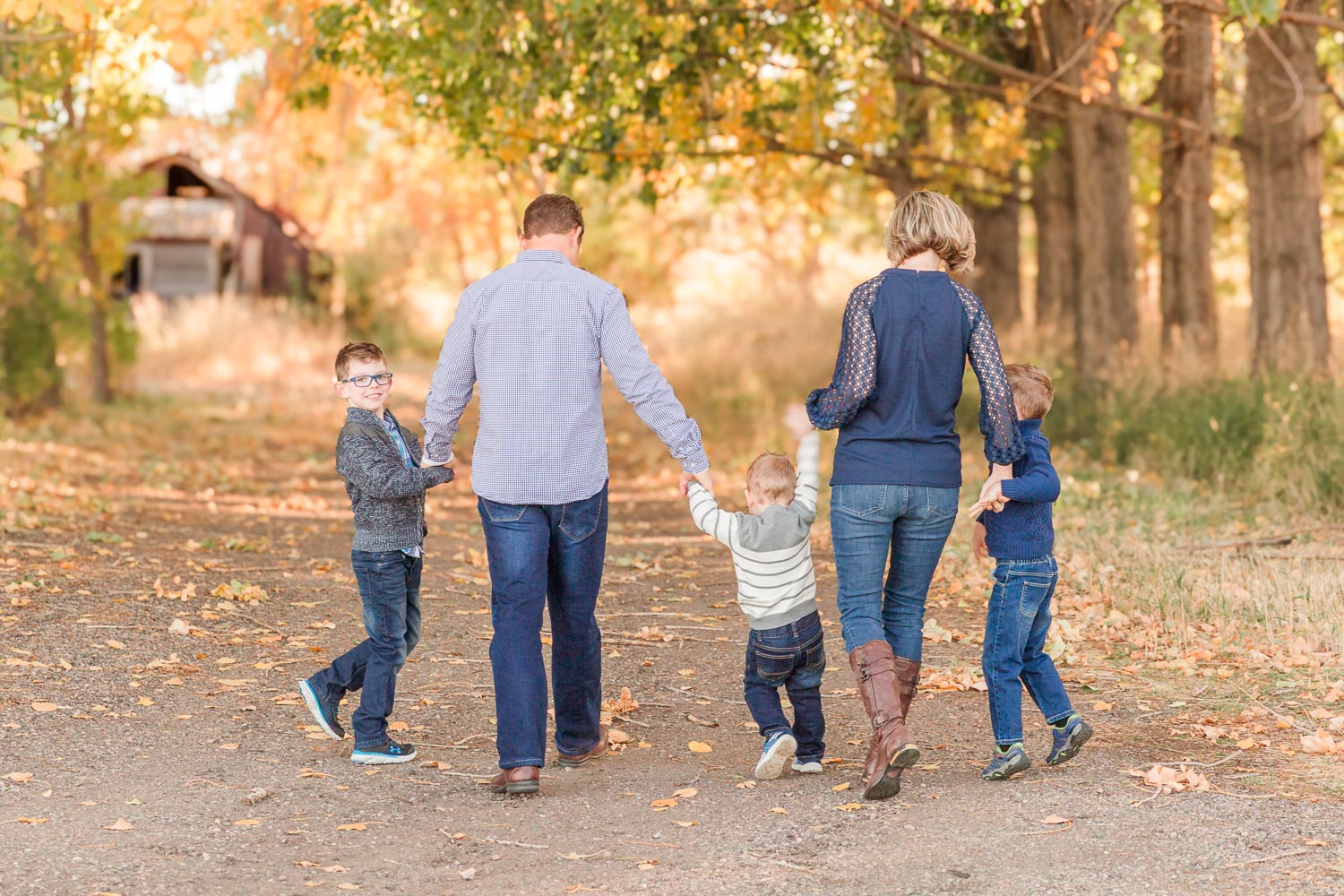 Fall family session in Erie, Colorado—parents and their three young children holding hands and walking through a tree-lined path with golden autumn leaves. Captured by Theresa Pelser, Erie family photographer specializing in joyful, candid moments.