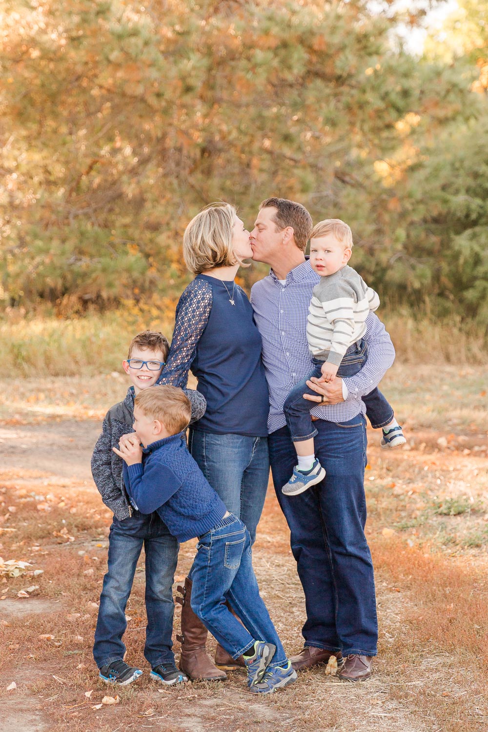 Family session at the Wise Homestead in Erie, Colorado—parents kissing while their three young sons cuddle and play around them during a golden fall afternoon. Captured by Theresa Pelser, Erie family photographer.