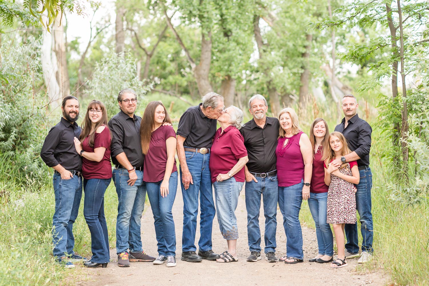 Extended family session at McKay Lake in Thornton, Colorado—four generations dressed in coordinated black and burgundy, smiling and standing together on a tree-lined path. Captured by Theresa Pelser, extended family photographer in Colorado.
