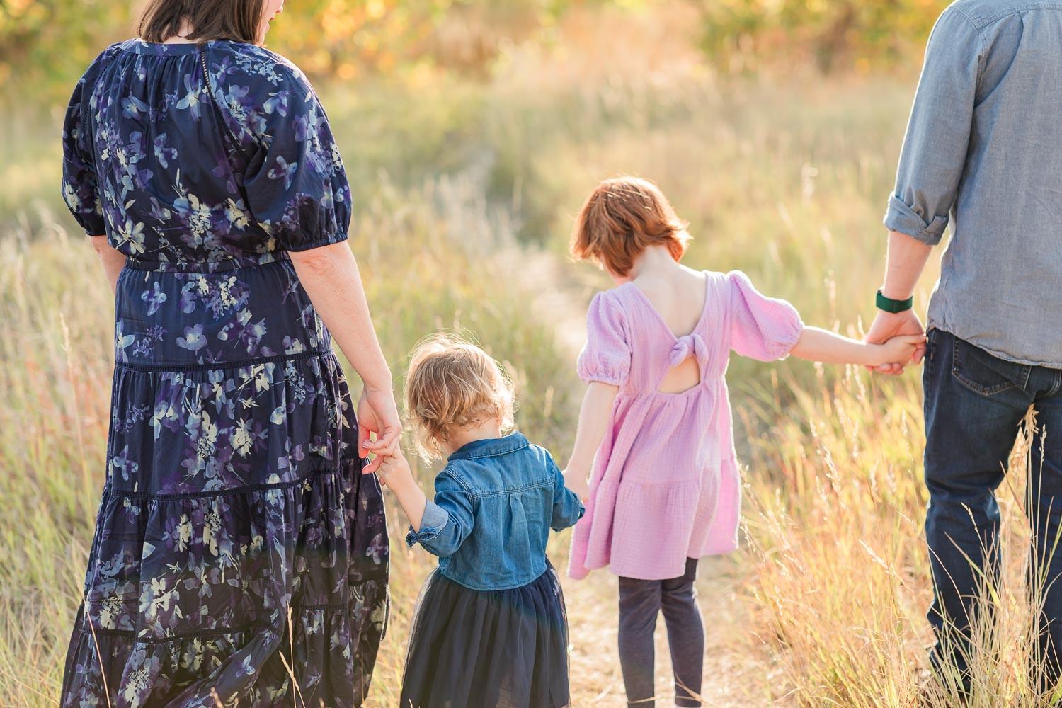 Family session at Coal Creek in Erie, Colorado—parents walking hand-in-hand with their two young daughters through a sunlit grassy field during fall. Captured by Theresa Pelser, Erie family photographer.