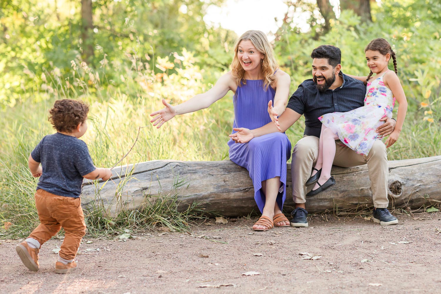 Family session at McKay Lake in Colorado—parents sitting on a fallen log with their daughter, reaching out with smiles as their toddler son runs toward them on a wooded trail. Captured by Theresa Pelser, lifestyle family photographer.