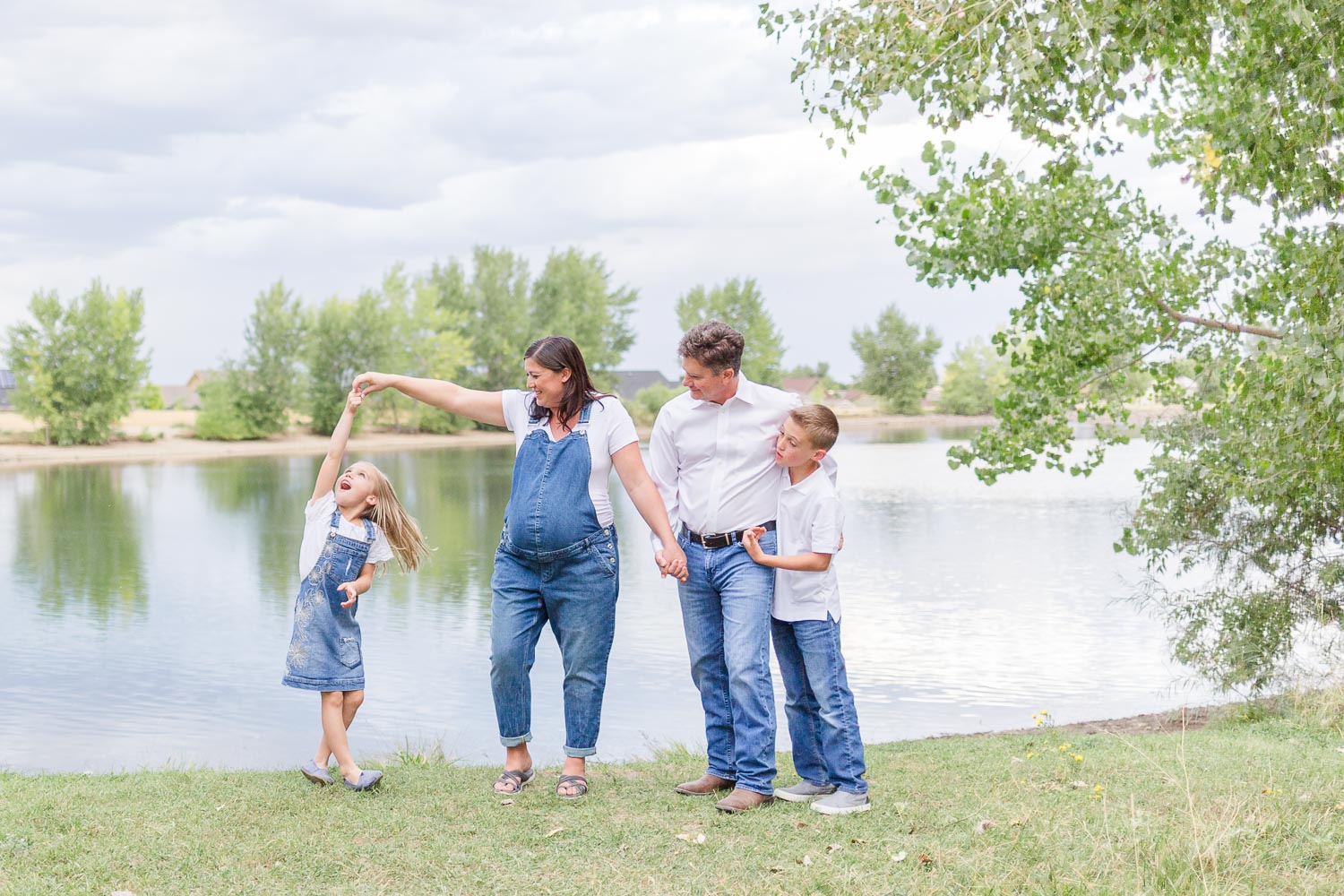 Maternity session at Thomas Reservoir in Erie, Colorado—expecting mother dancing with her young daughter while the father and son smile nearby, captured by the water on a cloudy day. Photographed by Theresa Pelser, Erie family and maternity photographer.