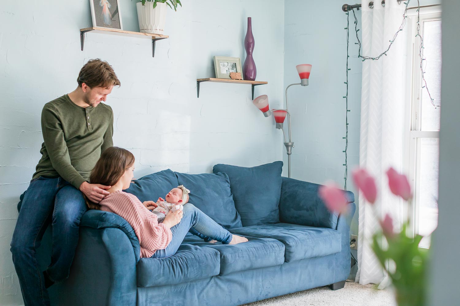 In-home newborn session in Broomfield, Colorado—parents sitting together on a blue couch, admiring their newborn baby in a cozy, light-filled living room. Captured by Theresa Pelser, Broomfield newborn and family photographer.