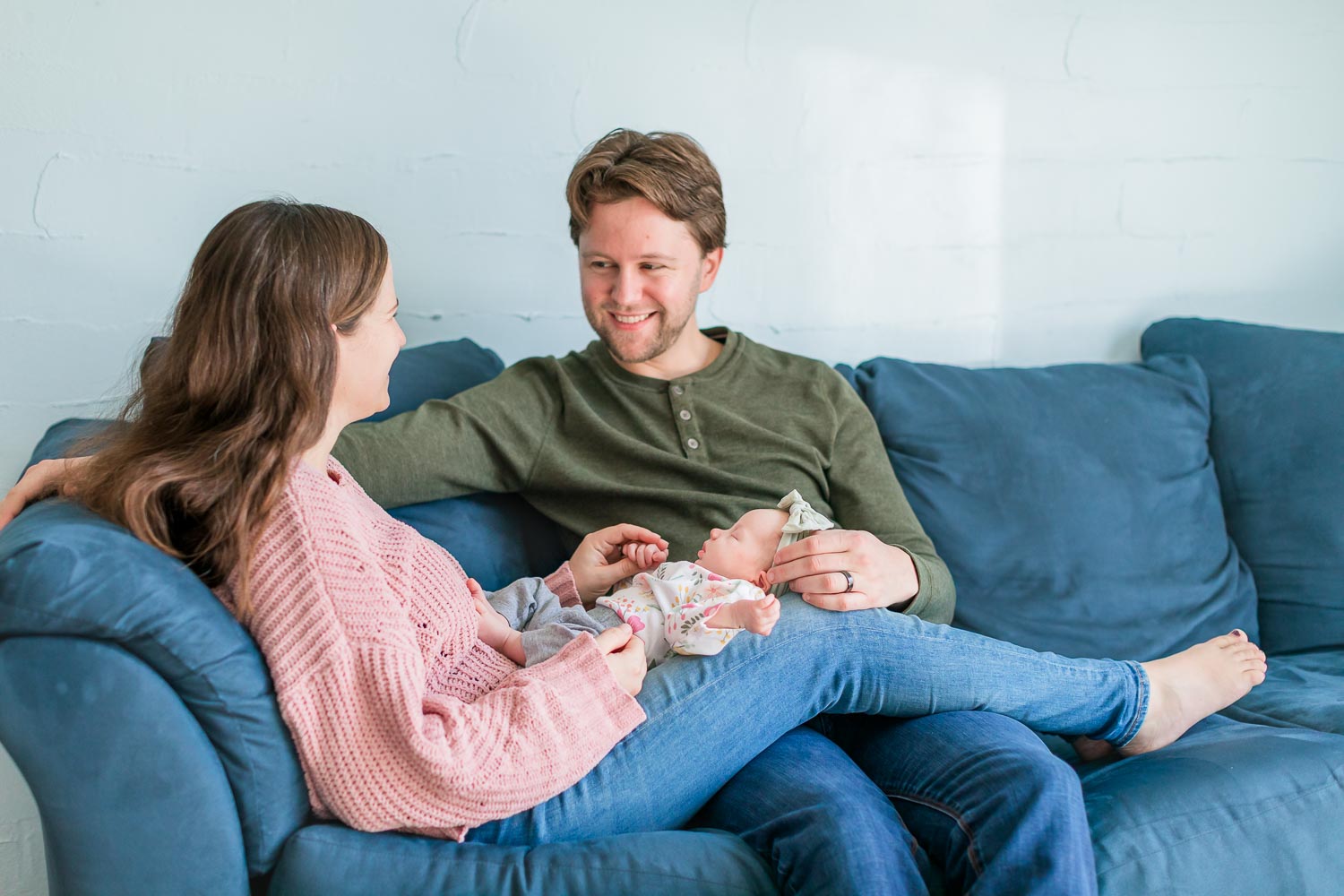 In-home newborn session in Broomfield, Colorado—parents sitting on a blue couch, smiling at each other while holding their newborn baby in a cozy, light-filled room. Captured by Theresa Pelser, Broomfield newborn and family photographer.
