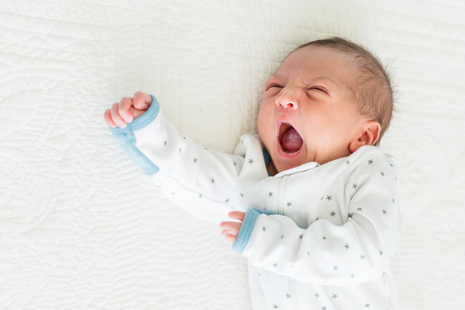 In-home newborn session in Erie, Colorado—newborn baby in a white onesie yawning while lying on a soft white bed. Captured by Theresa Pelser, Erie newborn and family photographer.