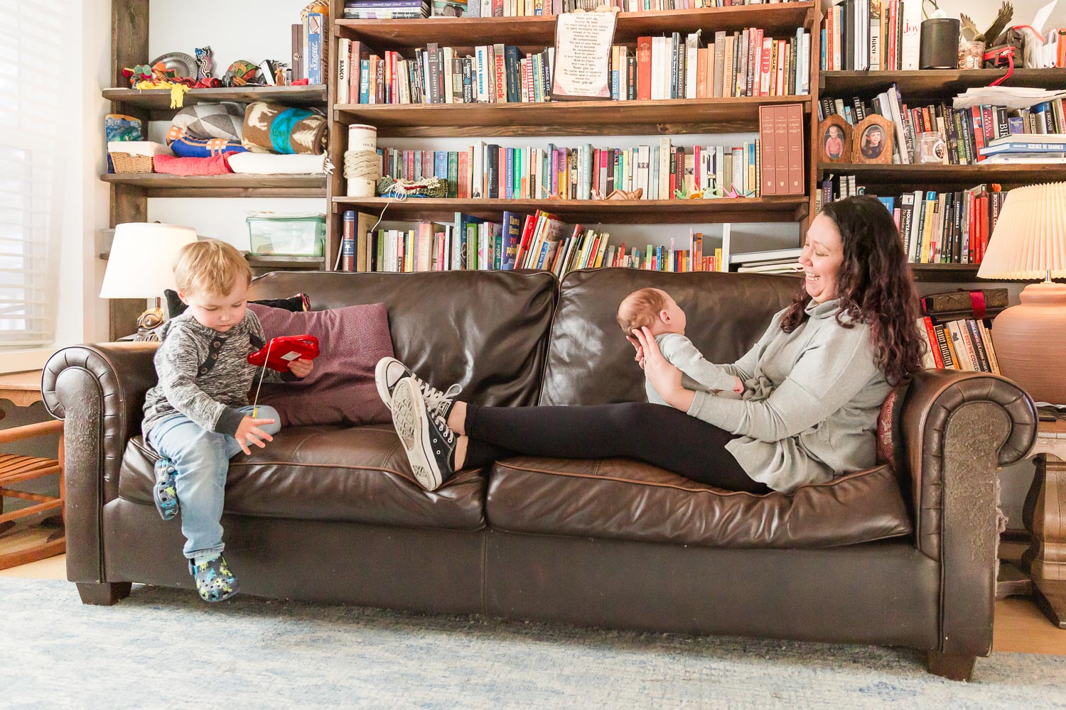 In-home newborn session in Boulder, Colorado—mother lying on a leather couch cradling her newborn while an older sibling plays nearby in a cozy living room with a full bookshelf. Captured by Theresa Pelser, Boulder newborn and family photographer.