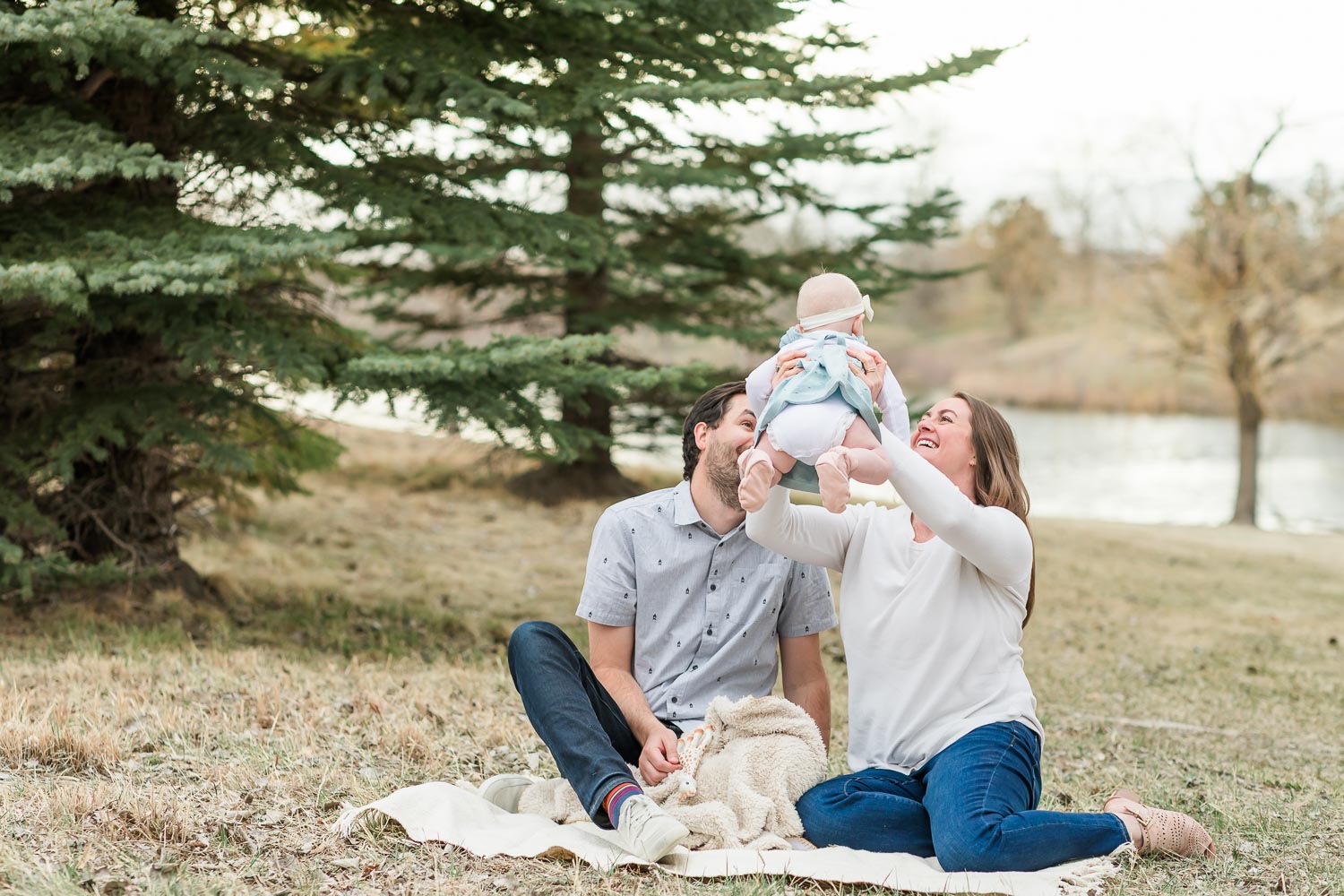 Outdoor newborn session at Anthem in Broomfield, Colorado—parents sitting on a blanket by the water as mom lifts their smiling baby into the air. Captured by Theresa Pelser, Broomfield newborn and family photographer.