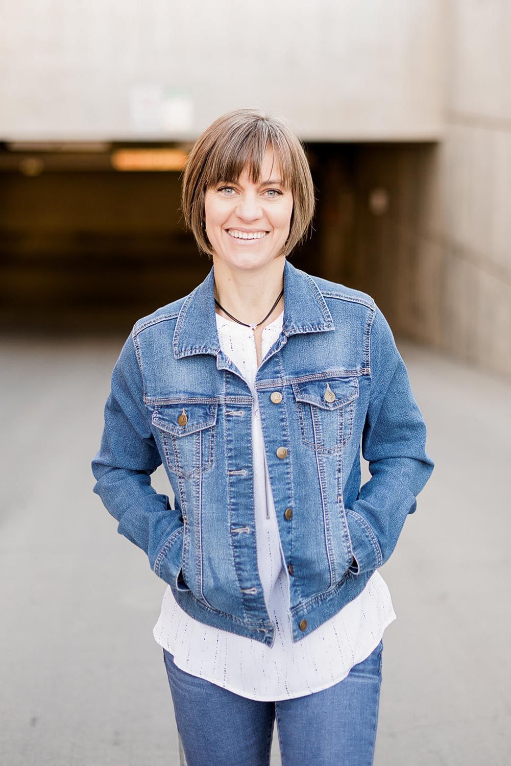 Theresa Pelser standing with hands in her pockets, smiling warmly in front of a neutral urban background—used in the welcome message on her homepage to reflect her laid-back, professional approach as an Erie family photographer.