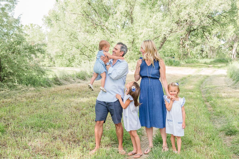 Family of five wearing shades of blue and white for summer family photos, baby dressed in romper, and little girls in matching white dresses with blue details.