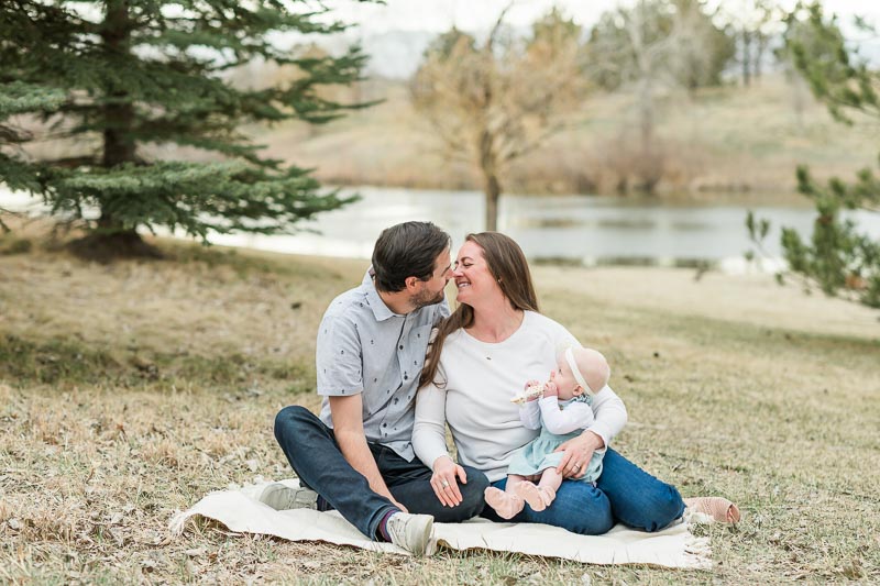 Parents wearing jeans holding baby dressed in light green romper for outdoor family photos.