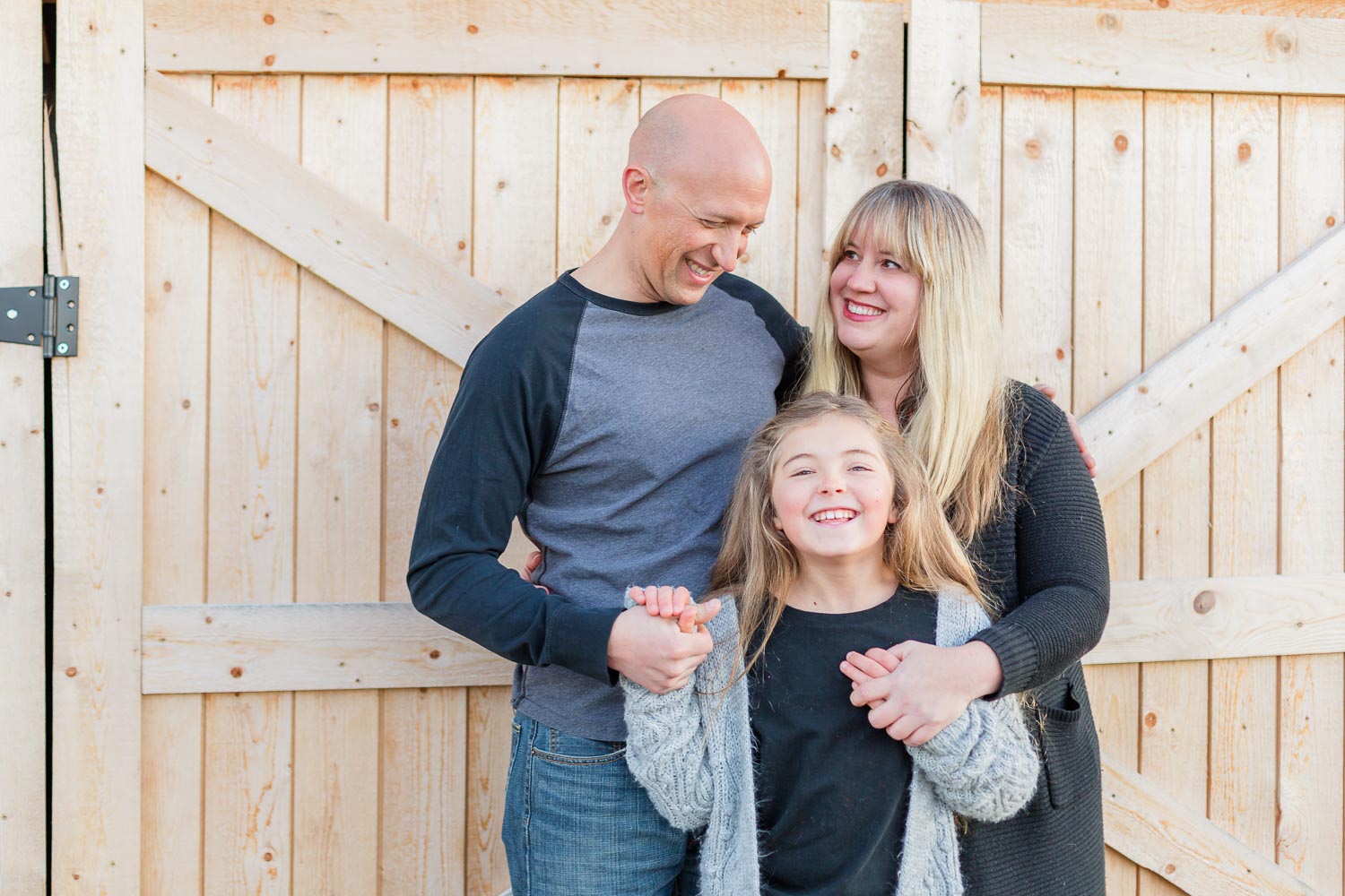 Smiling family of three standing in front of wooden barn doors during a fall photo session on their farm in Erie, Colorado.