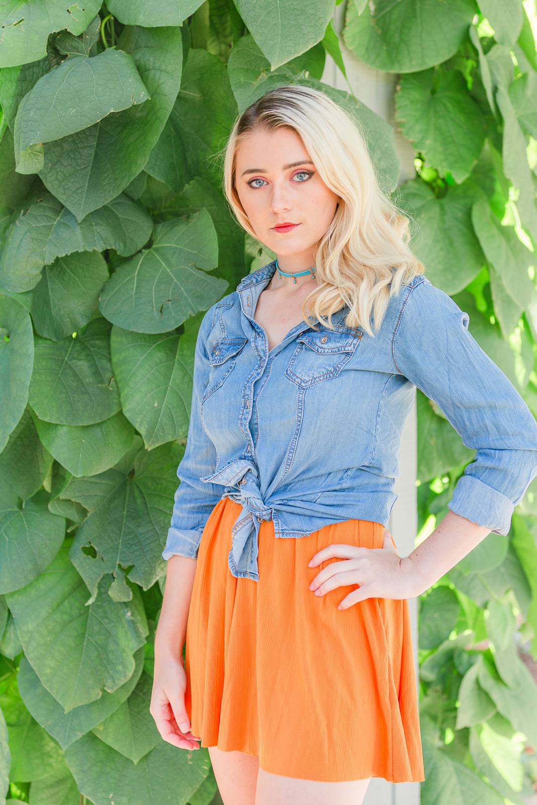 Photo of a senior girl wearing a denim shirt and orange mini skirt, paired with a statement necklace standing in front of a wall of large leaves.