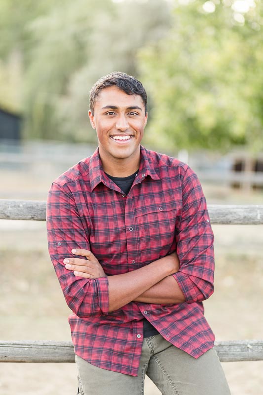 Senior photo of guy in a burgundy plaid shirt sitting casually on wooden fence with trees in the background.Senior photo of guy in a burgundy plaid shirt sitting casually on wooden fence with trees in the background.