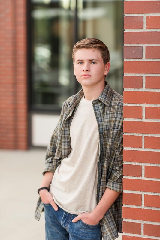 Senior photo of guy in neutral layers and subtle patterns leaning against a red brick wall in Erie, Colorado.