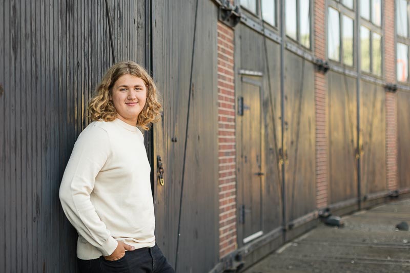 Senior photo of guy in black jeans and textured cream sweater leaning against rustic metal and wood doors at the Colorado Railroad museum.
