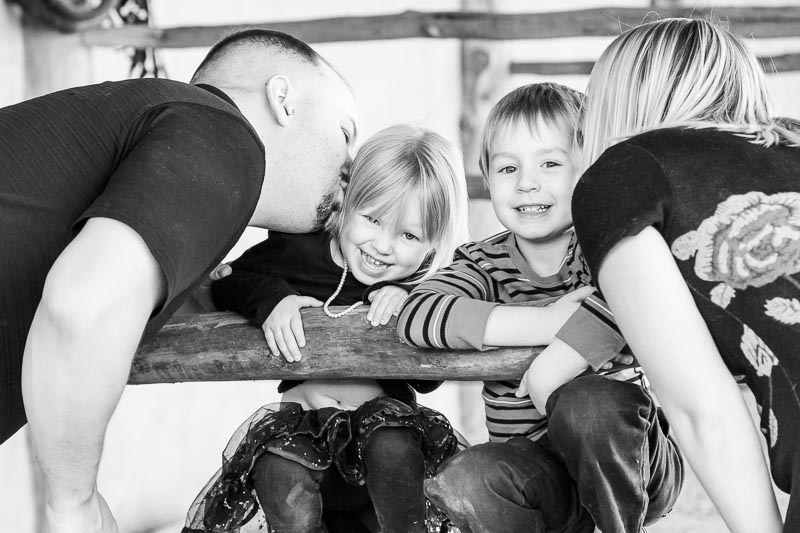 Mom and dad lean in to kiss their kids during an outdoor family photo session—candid laughter and connection.