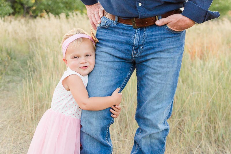 Shy toddler girl in a pink dress hugging her dad’s leg during outdoor family photos in Erie, Colorado.