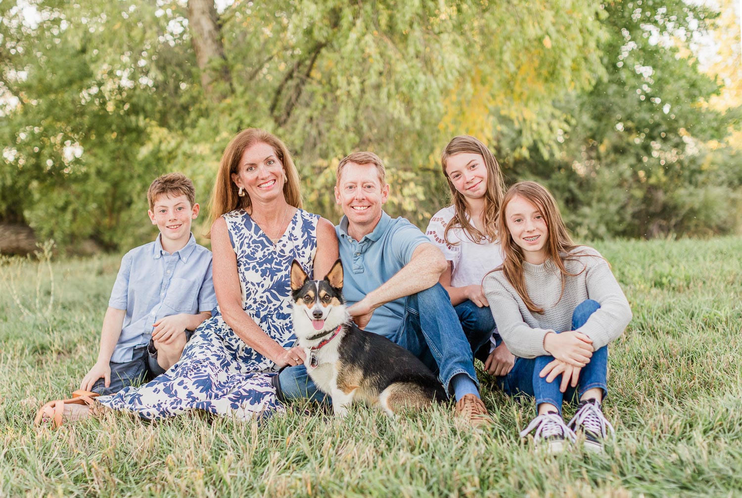 Fall mini session in Erie, Colorado—family of five with their dog sitting in a grassy field with trees in the background, sharing a relaxed and happy moment. Captured by Theresa Pelser, Erie family photographer.