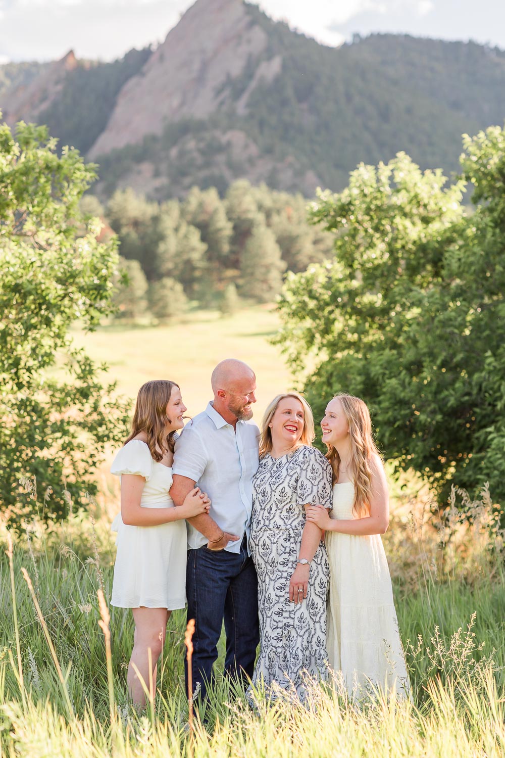 Outdoor family session in Boulder, Colorado with a family of four standing close together in a sunlit field near Chautauqua, relaxed and connected with mountain views behind them.