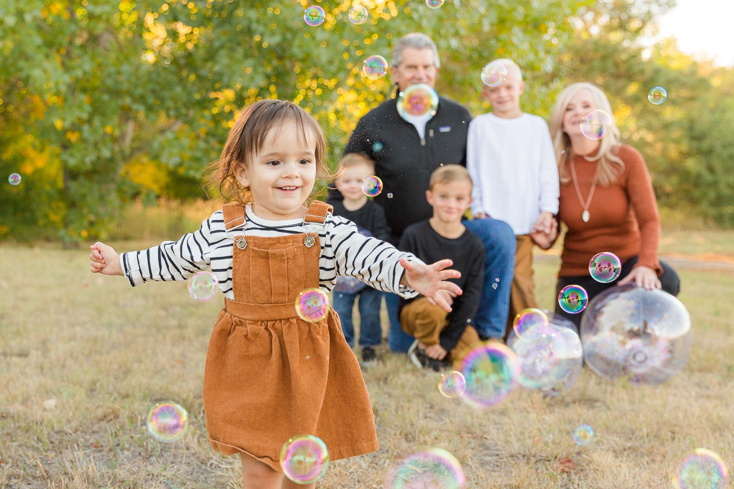 Extended family photo session with young child running toward bubbles while cousins and grandparents smile behind her in an open field near Erie, Colorado, natural and playful family moment