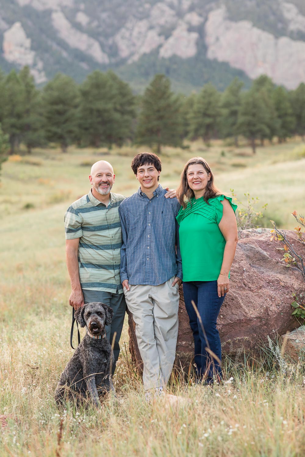 Family photography session in the Colorado foothills with parents, their teenage son, and black dog standing together in an open grassy field with mountain views behind them.