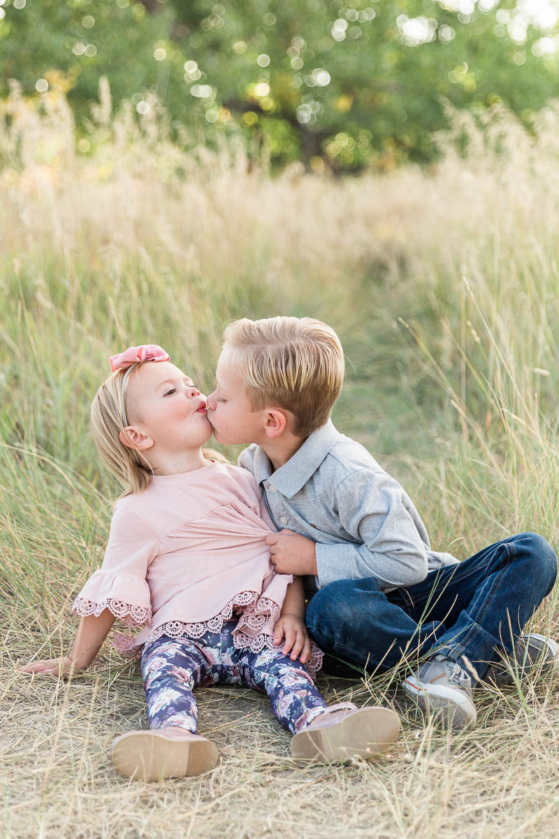 Young brother kissing his sister on the cheek as they sit together in tall grass.