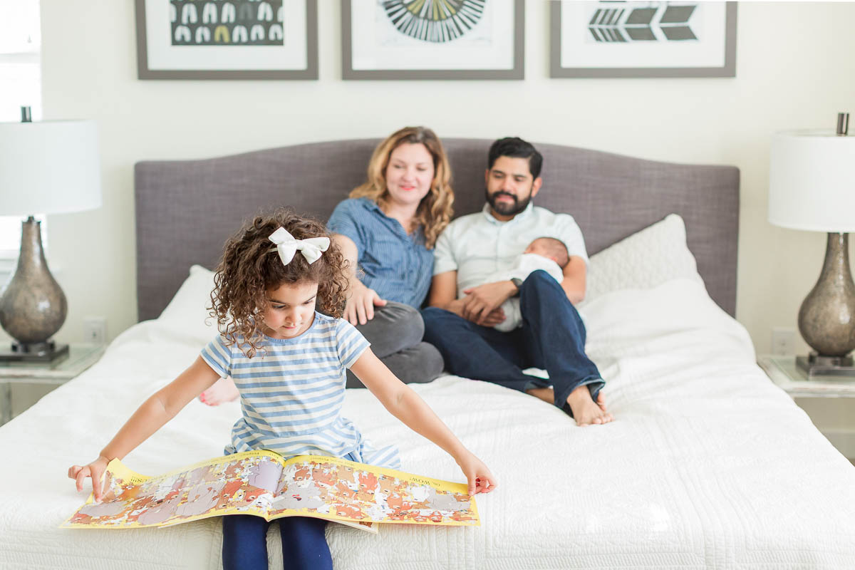 Older sister reads a book in the foreground while her parents sit on the bed with their new baby during an at-home newborn family session